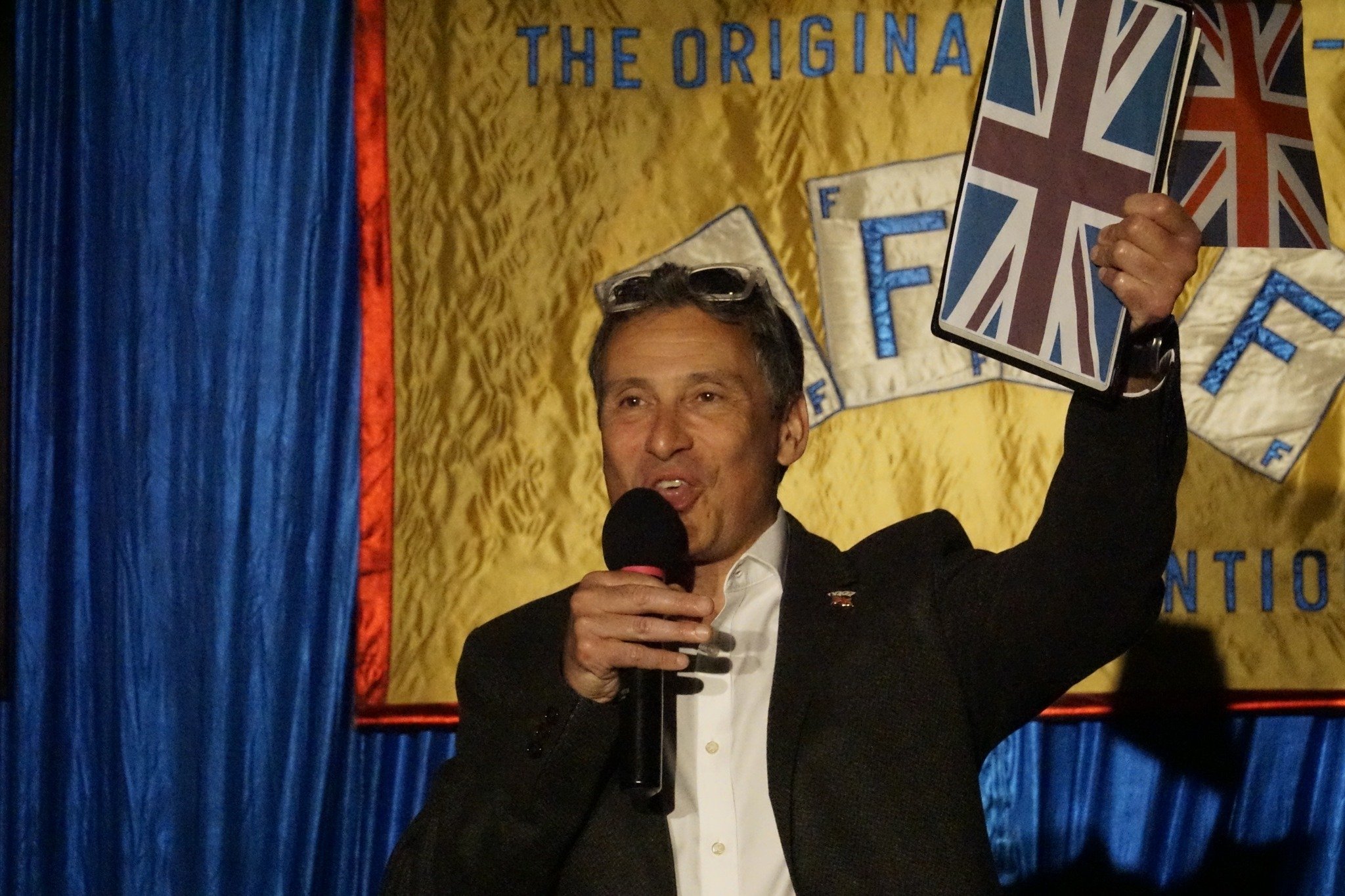 Darryl Rose Magician Performing On Stage, Holding A Microphone And A Union Jack Prop